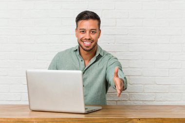 Genç Filipino adam oturan çalışma ile onun laptop streç el içinde kamera tebrik jest.