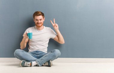 Genç kızıl saçlı öğrenci adam yerde oturan eğlenceli ve mutlu zafer bir jest yapıyor. Elinde bir kahve bardağı tutuyor..