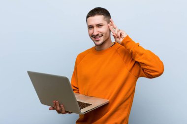 Zafer işaretini gösteren ve geniş bir şekilde gülümseyen bir laptop tutan genç beyaz adam.