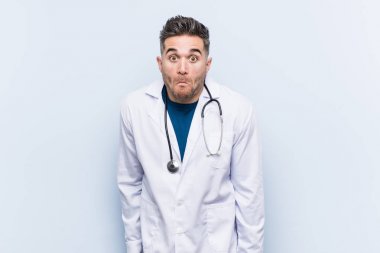 Young handsome doctor man shrugs shoulders and open eyes confused. clipart