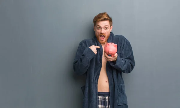 Young redhead man wearing pajama surprised, feels successful and prosperous. He is holding a piggy bank.