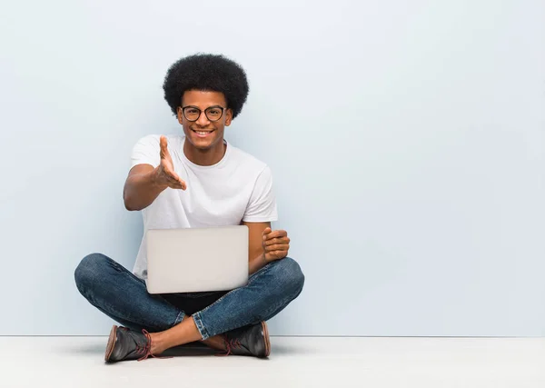 Young black man sitting on the floor with a laptop reaching out to greet someone