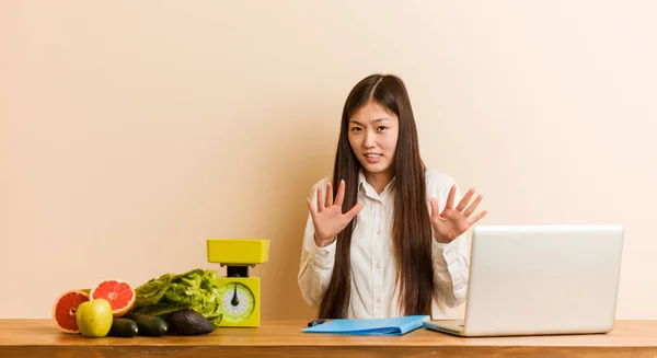 Young nutritionist chinese woman working with her laptop rejecting someone showing a gesture of disgust.