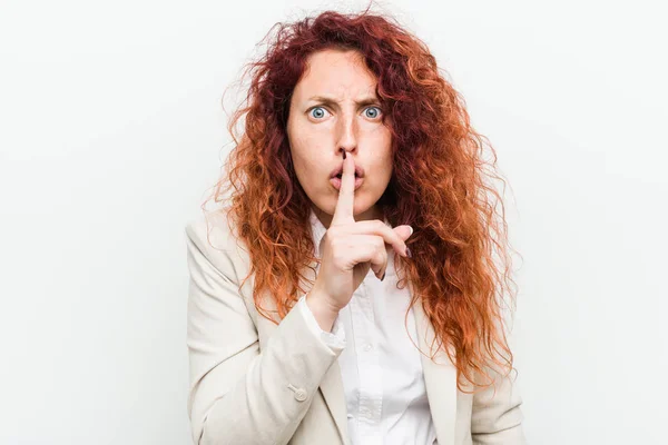 Young natural redhead business woman isolated against white background keeping a secret or asking for silence.