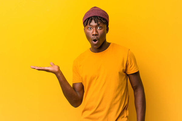 Young black man wearing rastas over yellow background impressed holding copy space on palm.