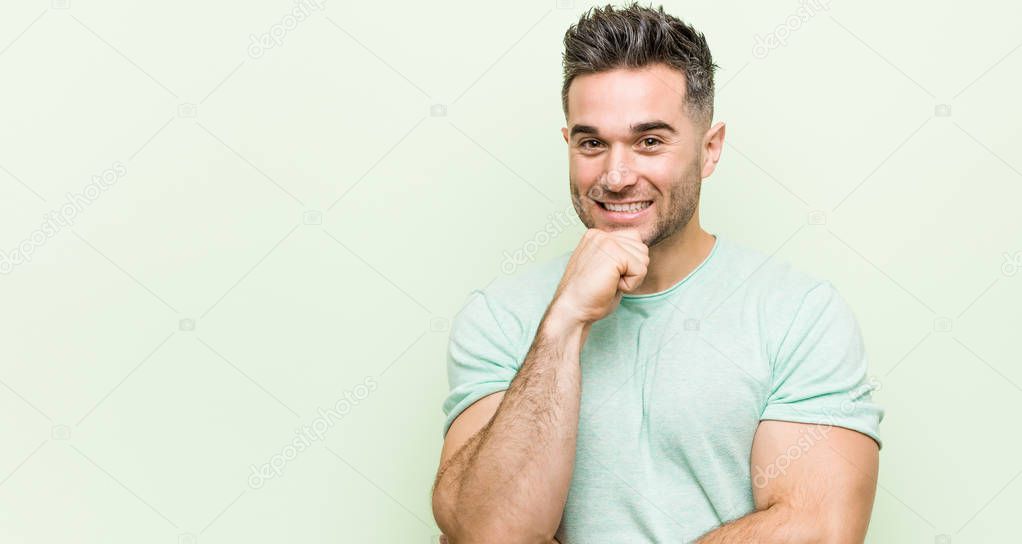 Young handsome man against a green background smiling happy and confident, touching chin with hand.
