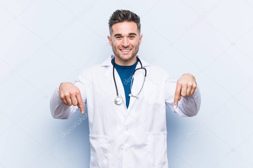 Young handsome doctor man points down with fingers, positive feeling.