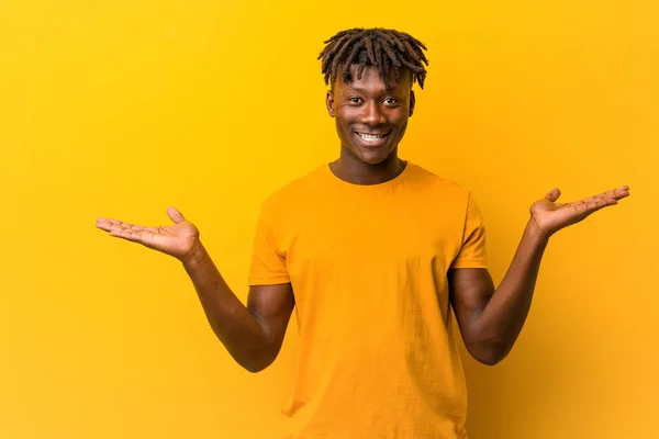 Young black man wearing rastas over yellow background makes scale with arms, feels happy and confident.