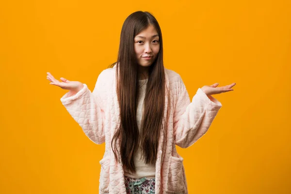 Young chinese woman in pajama doubting and shrugging him shoulders in questioning gesture.