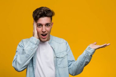 Denim gömlek giyen genç yakışıklı öğrenci bir avuç üzerinde kopya alanı tutar, yanak üzerinde el tutmak. Şaşırmış ve memnun.