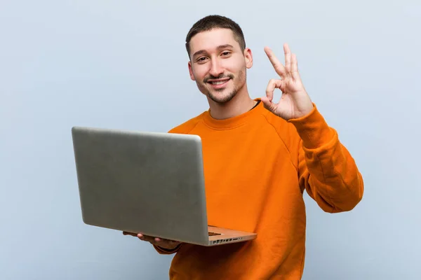 年轻的白人男子拿着笔记本电脑欢快和自信显示Ok手势 — 图库照片
