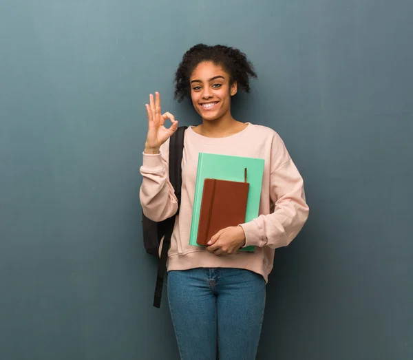 Young student black woman cheerful and confident doing ok gesture. She is holding books.
