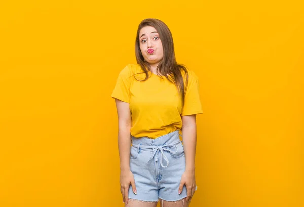 Young woman teenager wearing a yellow shirt blows cheeks, has tired expression. Facial expression concept.
