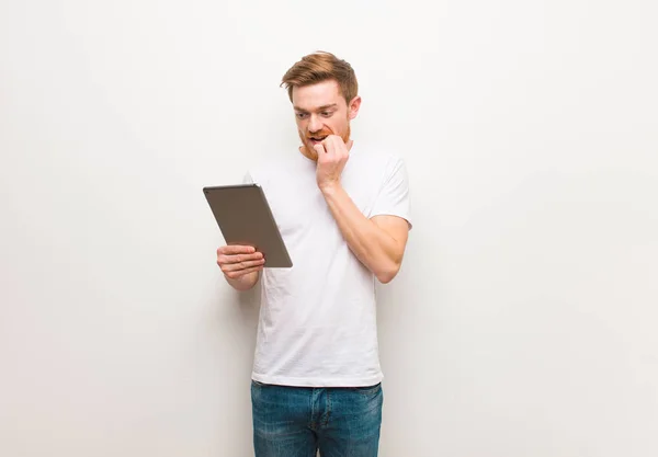 Young redhead man biting nails, nervous and very anxious. Holding a tablet.