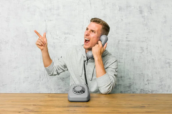 Young man talking on a vintage phone pointing with forefingers to a copy space, expressing excitement and desire.