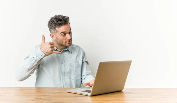 年轻的帅哥工作与他的笔记本电脑显示一个手机呼叫手势用手指 — 图库照片