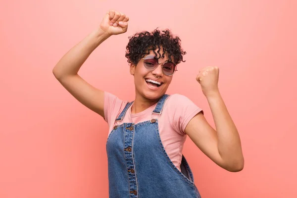 Young mixed african american teenager woman celebrating a special day, jumps and raise arms with energy.