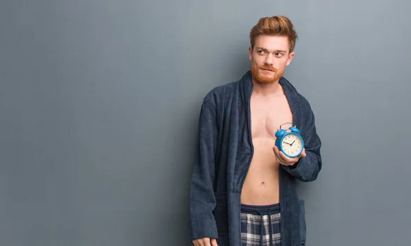 Young redhead man wearing pajama smiling confident and crossing arms, looking up. He is holding an alarm clock.