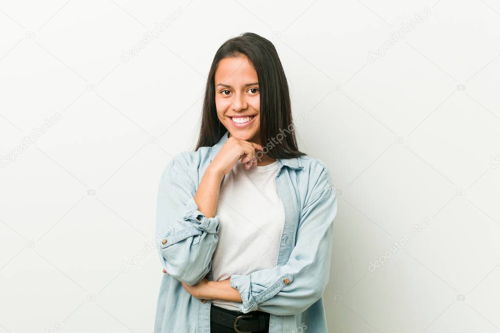 Young hispanic woman smiling happy and confident, touching chin with hand.