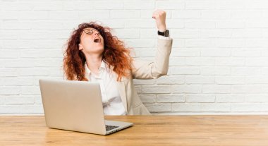 Genç kızıl saçlı kıvırcık kadın bir zaferden sonra yumruk yükselterek dizüstü bilgisayar ile çalışan, kazanan kavram.