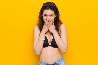 Genç esmer kadın el ile ağız kapsayan, bir şey hakkında gülüyor sarı arka plana karşı bir bikini giyiyor.
