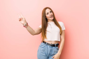 Pembe bir duvara karşı genç kızıl saçlı zencefil kadın neşeli ve kaygısız parmakları ile bir barış sembolü gösteren.