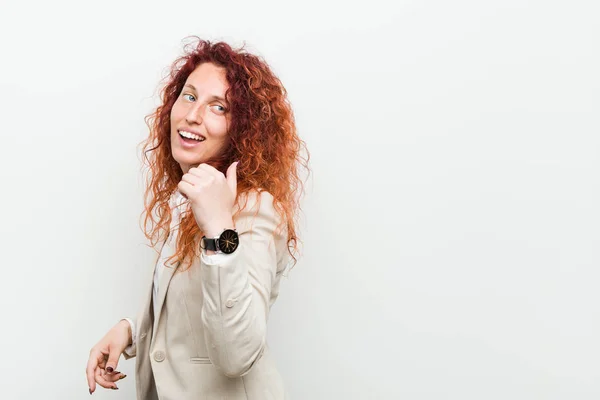 Young natural redhead business woman isolated against white background points with thumb finger away, laughing and carefree.