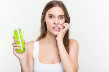 Aloe vera şişesi tutan genç beyaz kadın tırnaklarını ısırıyor, sinirli ve çok endişeli.
