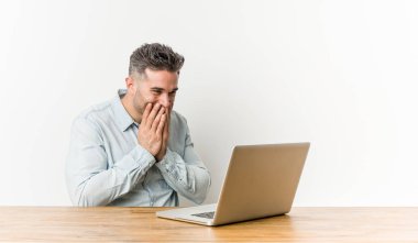 Dizüstü bilgisayarıyla çalışan genç yakışıklı adam bir şeye gülüyor, ağzı elleriyle kapatıyor..