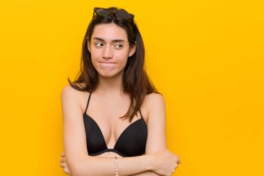 Genç esmer kadın sarı arka plan karşı bir bikini giyen karışık, şüpheli ve emin hissediyor.