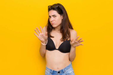 Genç esmer kadın iğrenme bir jest gösteren birini reddederek sarı arka plan karşı bir bikini giyiyor.