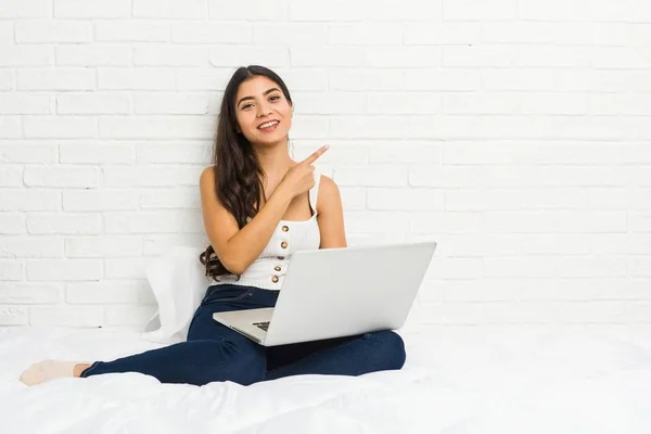 ベッドの上でノートパソコンを持って作業している若いアラブ人女性は 笑顔で脇を指差し 空白のスペースで何かを示しています — ストック写真