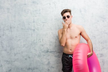 Pembe şişme çörek tutan genç adam elinde ağız kapsayan yorgun bir jest gösteren esneme bir mayo giyen.