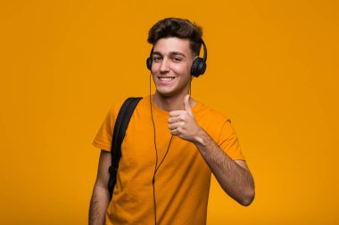 Genç serin öğrenci adam zafer işareti gösteren kulaklık ile müzik dinlerken ve geniş gülümseyerek.