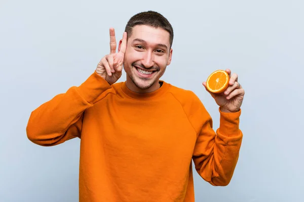 Joven Hombre Caucásico Sosteniendo Una Naranja Mostrando Signo Victoria Sonriendo — Foto de Stock