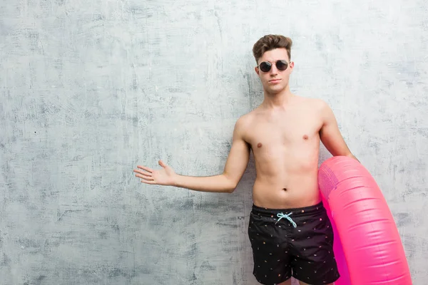 Pembe şişme çörek tutan genç adam hoş bir ifade gösteren bir mayo giyiyor.