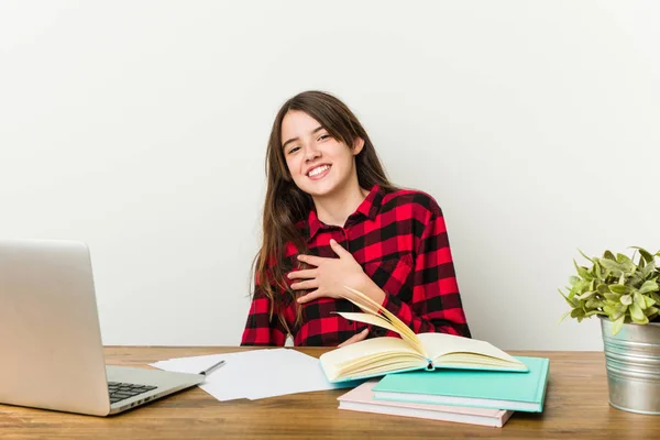 Young teenager going back to her routine doing homeworks laughs happily and has fun keeping hands on stomach.