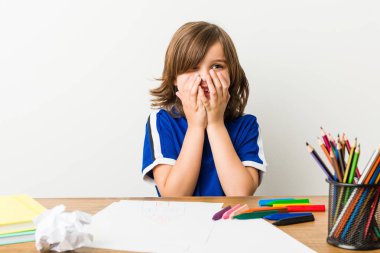 Küçük çocuk resim yapıyor ve masasında ödevler yapıyor bir şeye gülüyor, ağzı elleriyle kapatıyor..