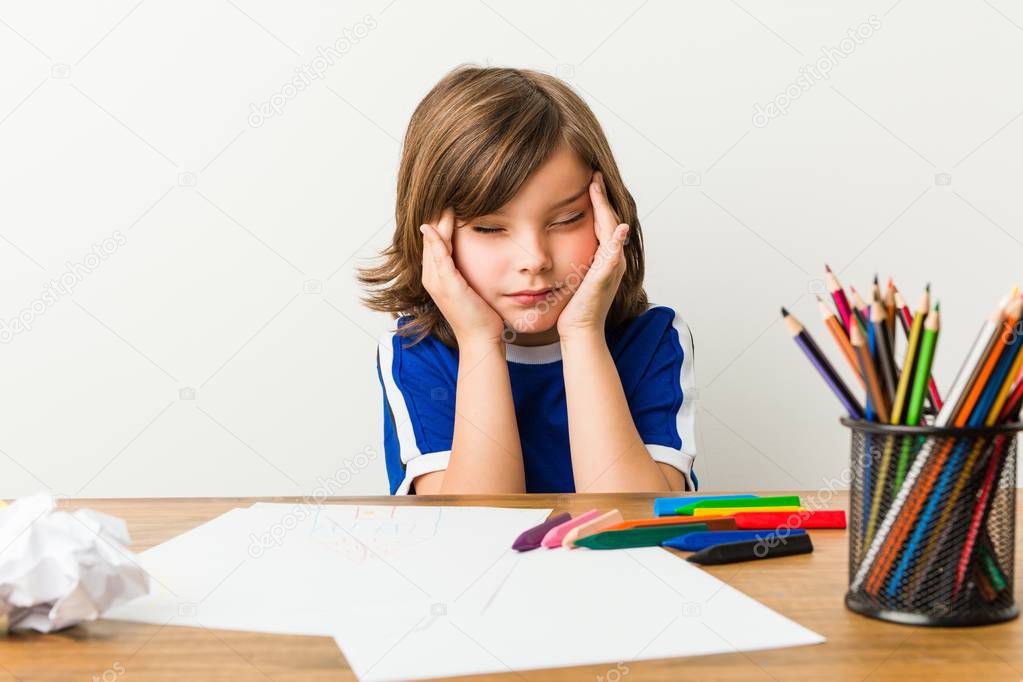 Little boy painting and doing homeworks on his desk touching temples and having headache.