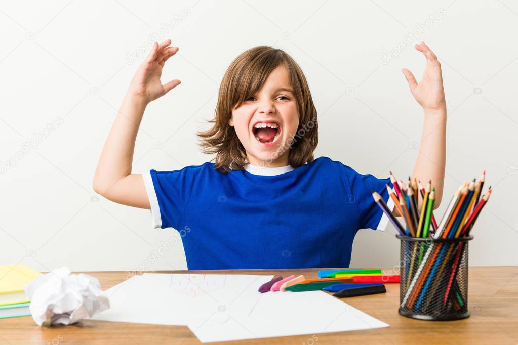 Little boy painting and doing homeworks on his desk receiving a pleasant surprise, excited and raising hands.