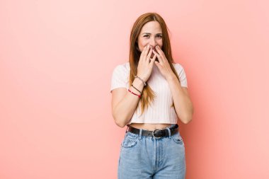 Pembe bir duvara karşı genç kızıl saçlı zencefil kadın bir şey hakkında gülüyor, elleriyle ağız kapsayan.