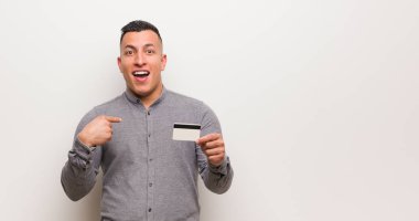Young latin man holding a credit card surprised, feels successful and prosperous clipart