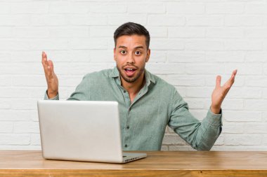 Genç filipinli adam onun dizüstü bilgisayar ile çalışan oturan hoş bir sürpriz, heyecanlı ve ellerini yükselterek alma.