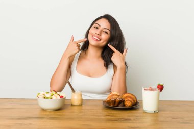 Genç kıvrımlı kadın bir kahvaltı gülümsüyor alarak, ağızda parmaklarını işaret.