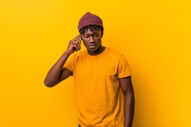 Şapka takarak sarı bir arka plan aleyhine duran ve telefon kullanan afrikalı genç adam