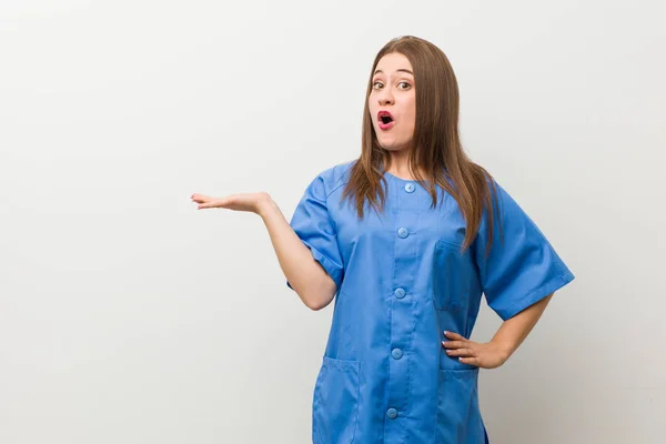 Junge Krankenschwester Vor Weißer Wand Beeindruckt Mit Kopierraum Auf Handfläche — Stockfoto