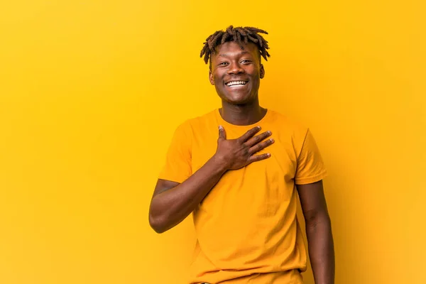 Young black man wearing rastas over yellow background laughs out loudly keeping hand on chest.