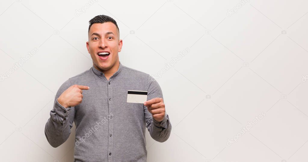 Young latin man holding a credit card surprised, feels successful and prosperous