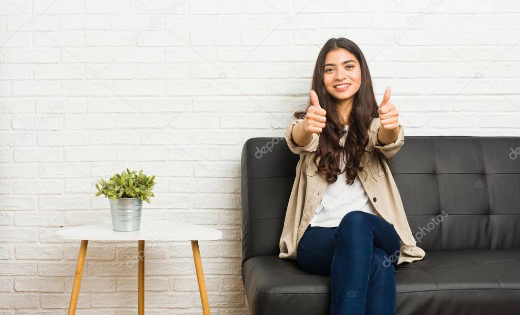 Young arab woman sitting on the sofa with thumbs ups, cheers about something, support and respect concept.