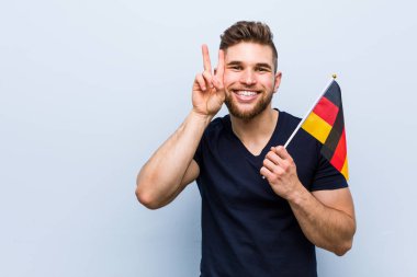 Zafer işareti gösteren ve geniş gülümseyen bir Almanya bayrağı tutan genç beyaz adam.